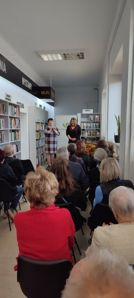 na zdjęciu sala biblioteczna wypełniona osobami, które przeszły na spotkanie autorskie z Joanną Jax. Z samego przodu dyrektor biblioteki wita autorkę obie stoją.