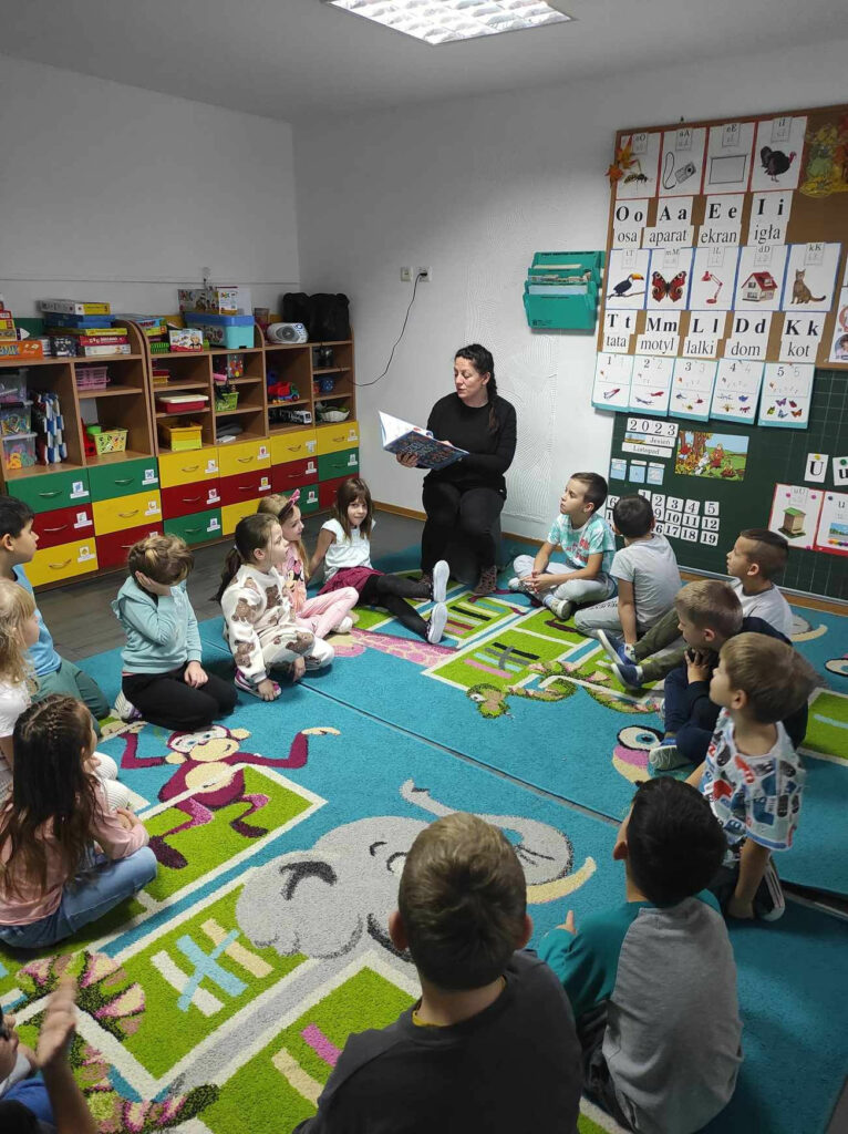 na zdjęciu widać salę przedszkolną, dzieci siedzą w kole na dywanie, bibliotekarz czyta książkę