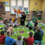 na zdjęciu widać sale przedszkolną, dzieci siedzą w kole na dywanie, bibliotekarz czyta książkę