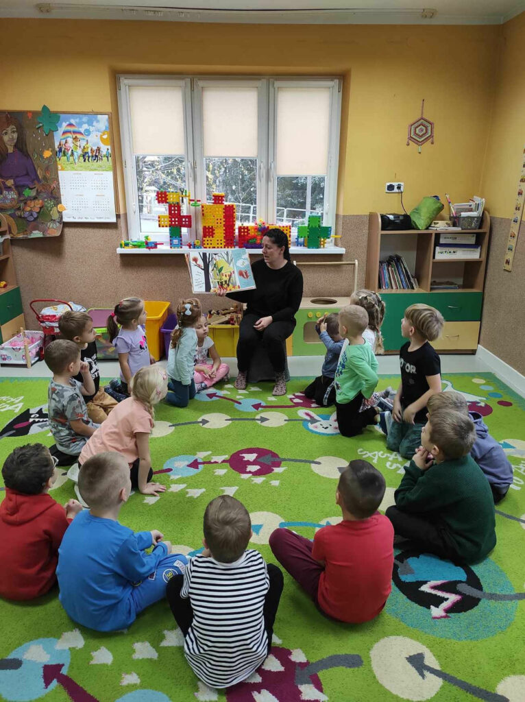 na zdjęciu widać sale przedszkolną, dzieci siedzą w kole na dywanie, bibliotekarz czyta książkę