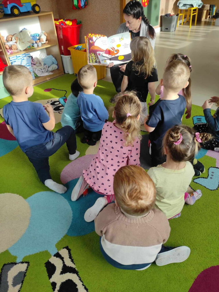 na zdjęciu widać salę przedszkolną, dzieci siedzą na dywanie, bibliotekarz czyta książkę