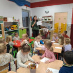Na zdjęciu dzieci siedza przy stolikach przed sobą mają kartki i kredki, którymi rysują. Pani bibliotekarka stoi i pokazuje rysunek dzieciom.
