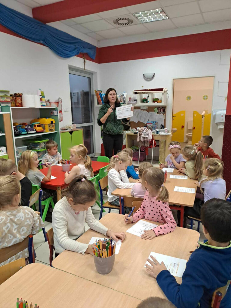 Na zdjęciu dzieci siedza przy stolikach przed sobą mają kartki i kredki, którymi rysują. Pani bibliotekarka stoi i pokazuje rysunek dzieciom.