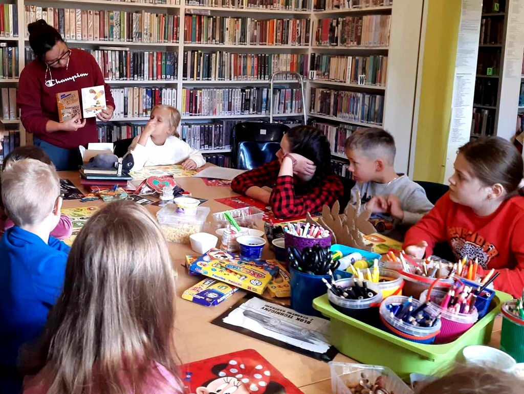 na zdjęciu widać dzieci siedzące przy stole w sali bibliotecznej oraz bibliotekarkę pokazująca im obrazki