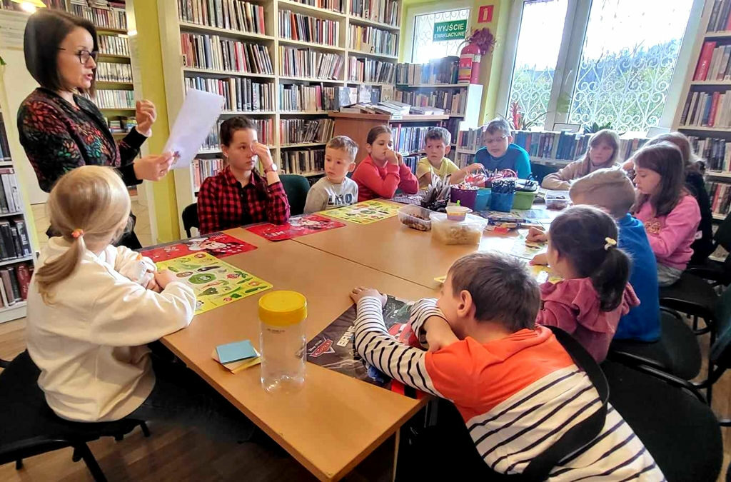 na zdjęciu widać dzieci siedzące przy stole w sali bibliotecznej oraz bibliotekarkę która im czyta