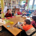 na zdjęciu widać dzieci siedzące przy stole w sali bibliotecznej oraz bibliotekarkę która im czyta