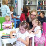 W sali bibliotecznej przy stolikach siedzą rodzice wraz z najmłodszymi dziećmi i wykonują prace plastyczne