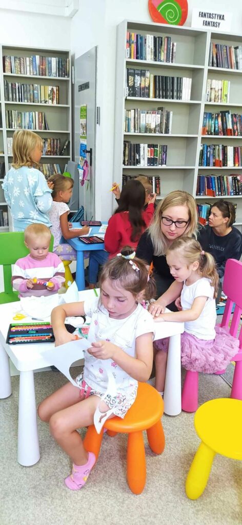 W sali bibliotecznej przy stolikach siedzą rodzice wraz z najmłodszymi dziećmi i wykonują prace plastyczne