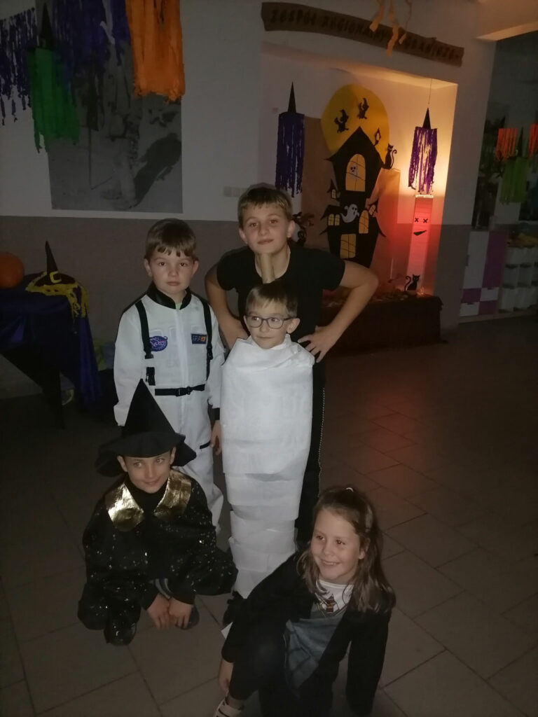 Na zdjęciu widocznych jest 5 dzieci (4 chłopców i 1 dziewczynka). Jeden z chłopców owinięty jest papierem toaletowym na podobieństwo mumii. W tle dekoracje tematyczne.