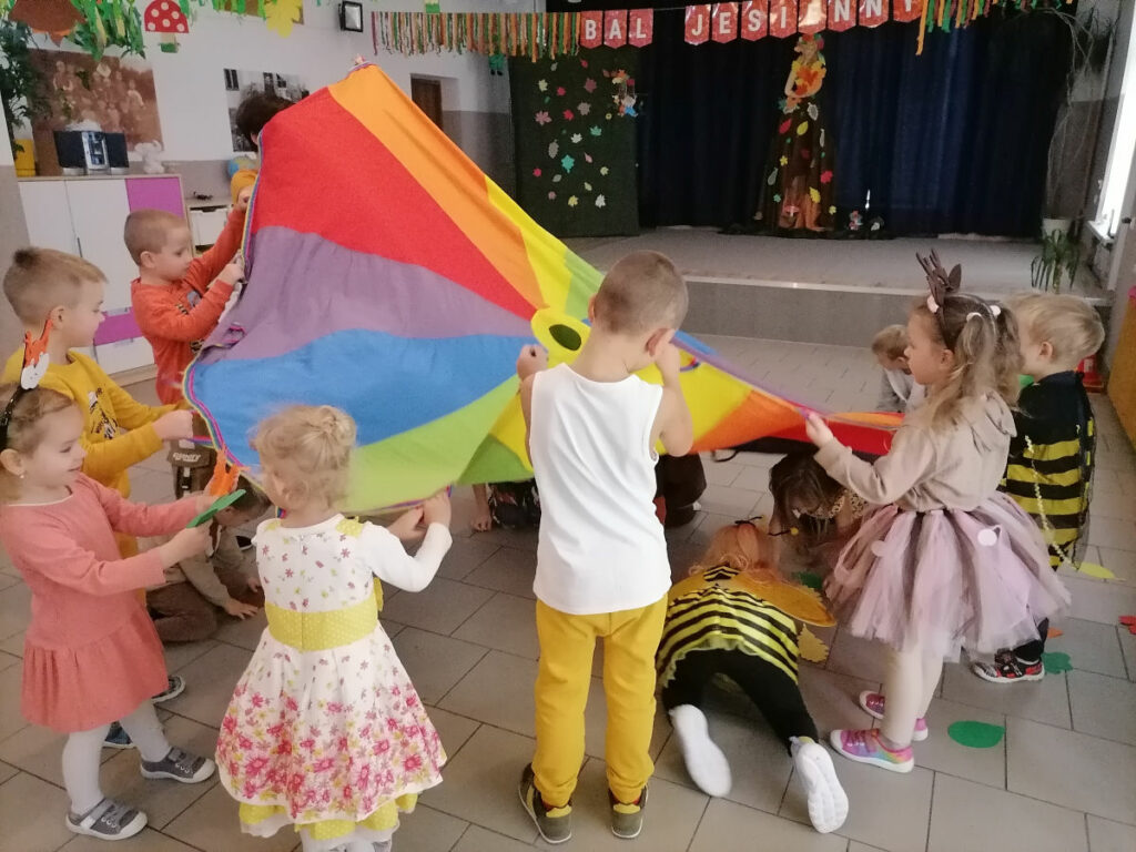 Na zdjęciu widoczna jest grupa dzieci w kolorowych strojach biorących udział w zabawach z chustą animacyjną.
