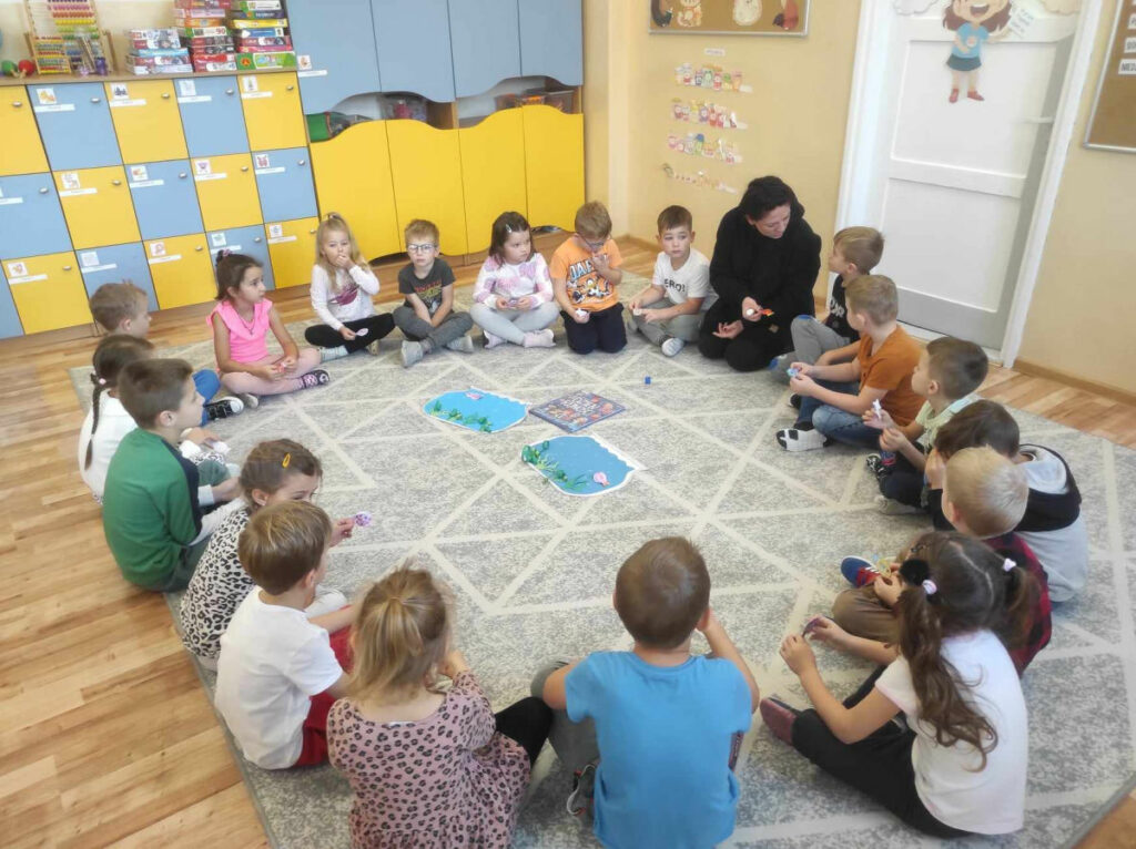 na zdjęciu widać salę przedszkolną, dzieci siedzą w kole na dywanie, wykonują wspólną pracę plastyczną