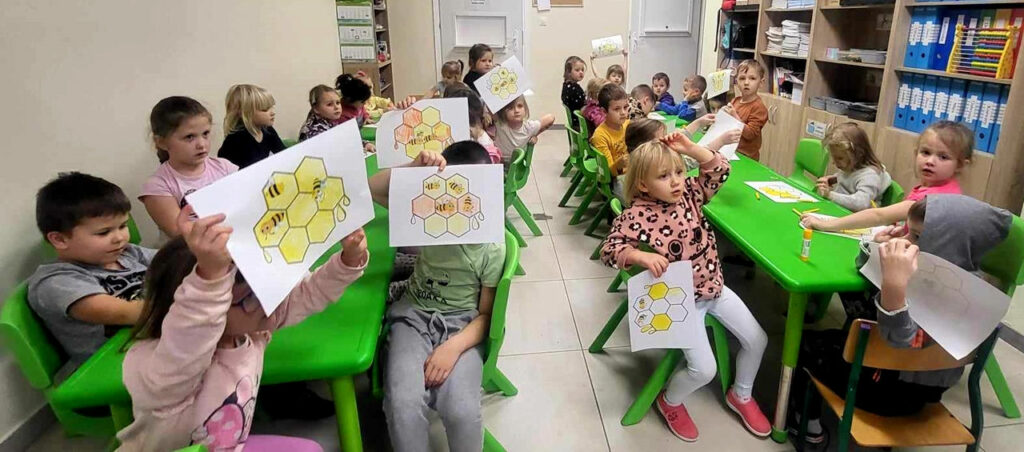 na zdjęciu widać dzieci siedzące przy zielonych stolikach i trzymające przez siebie pomalowane obrazki