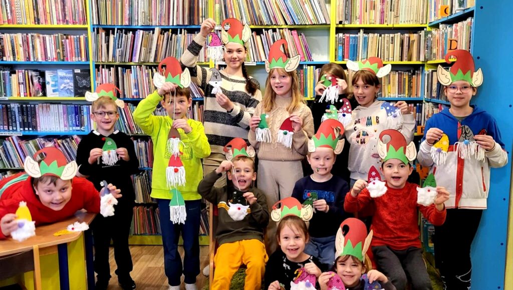 Zdjęcie przedstawia dzieci w czapkach elfów stojące na tle regału z książkami