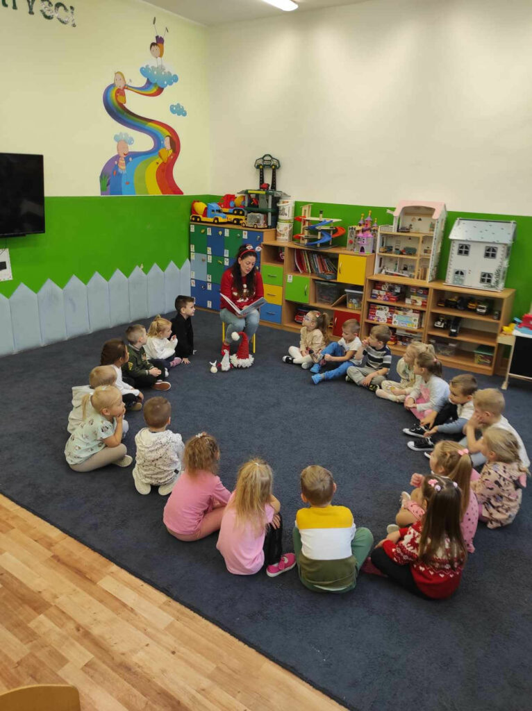 na zdjęciu widać sale przedszkolną, dzieci siedzą w kole na dywanie, bibliotekarz czyta książkę