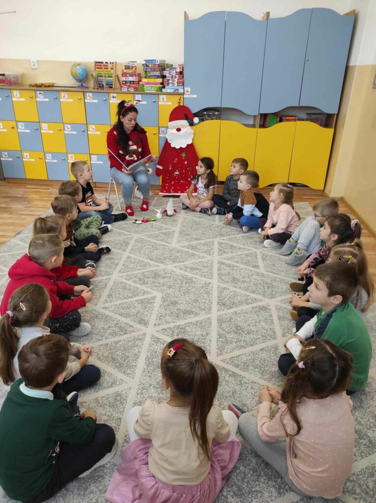 na zdjęciu widać salą przedszkolną, dzieci siedzą w kole na dywanie, bibliotekarz czyta książkę