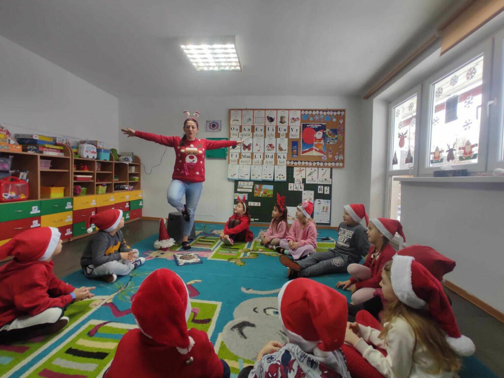 na zdjęciu widać salę przedszkolną, na dywanie siedzą dzieci, bibliotekarz prezentuje zabawę