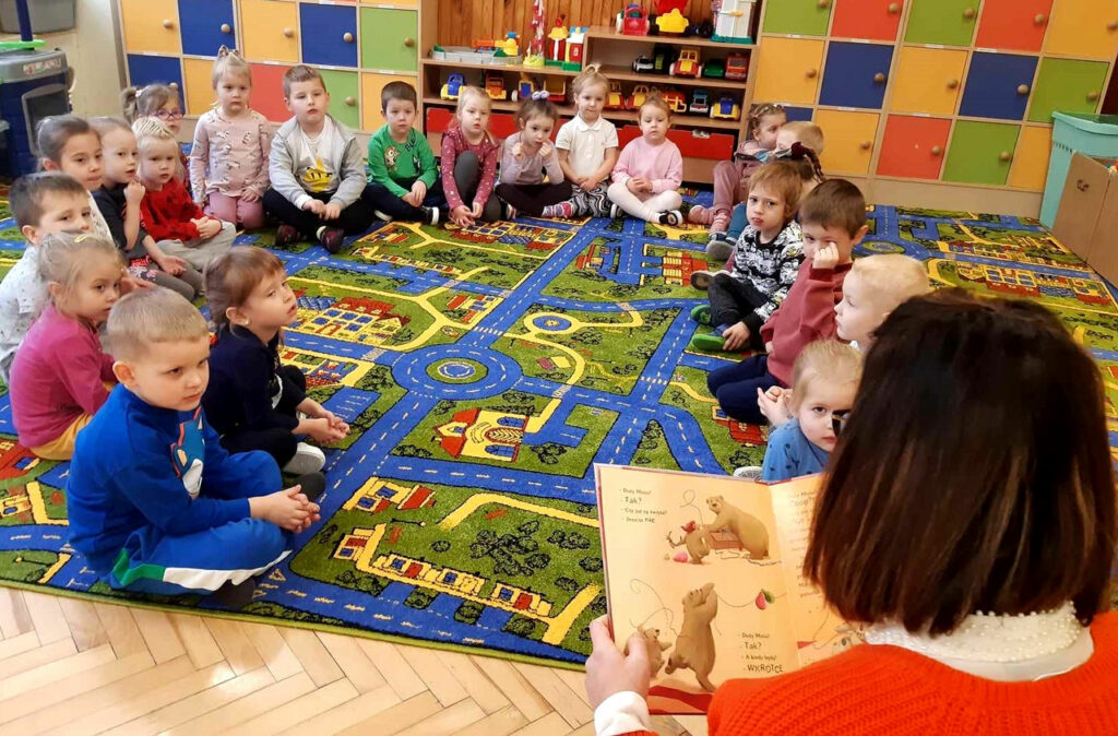 na zdjęciu widać dzieci siedzące na kolorowym dywanie w sali przedszkolnej