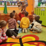 na zdjęciu widać dzieci na dywanie i czytającą im bibliotekarkę