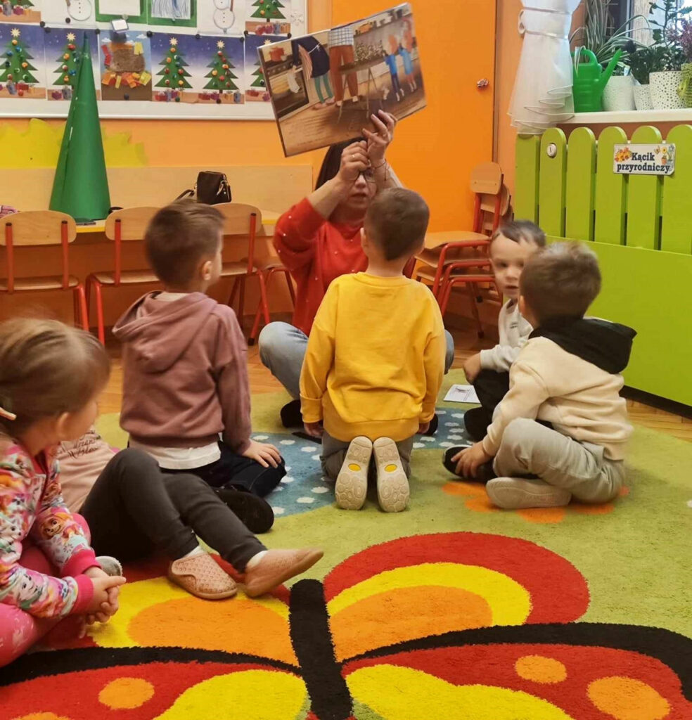na zdjęciu widać dzieci na dywanie i czytającą im bibliotekarkę