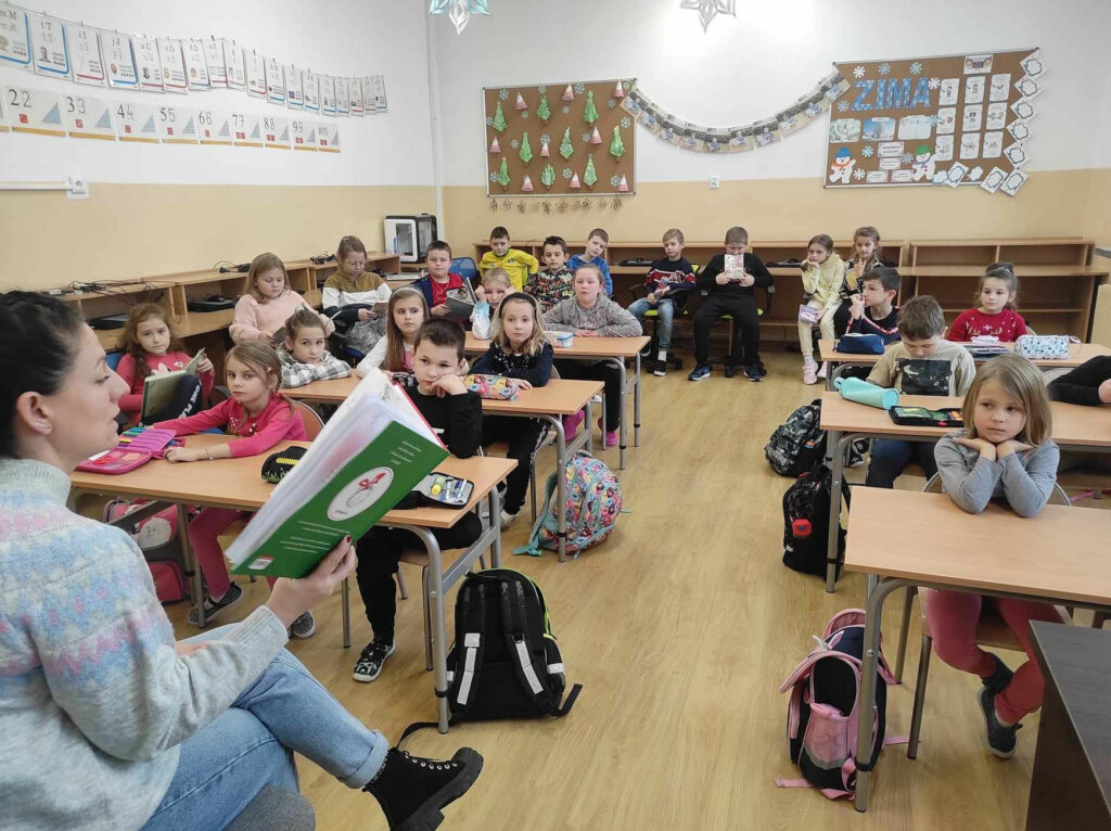 na zdjęciu widać salę szkolną, dzieci siedzą przy stolikach, bibliotekarz czyta książkę