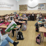 na zdjęciu widać salę szkolną, dzieci siedzą przy stolikach, bibliotekarz czyta książkę