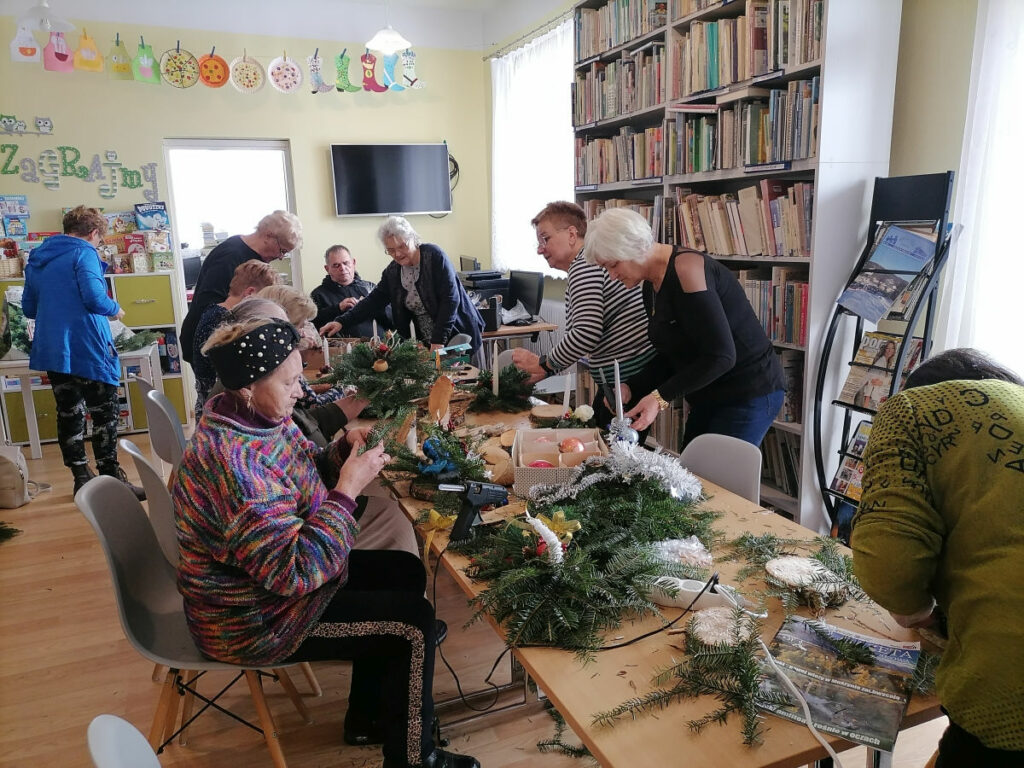 Na zdjęciu widoczna jest 1 pan i grupa 10 pań wykonujących stroiki bożonarodzeniowe. W tle regały z książkami.