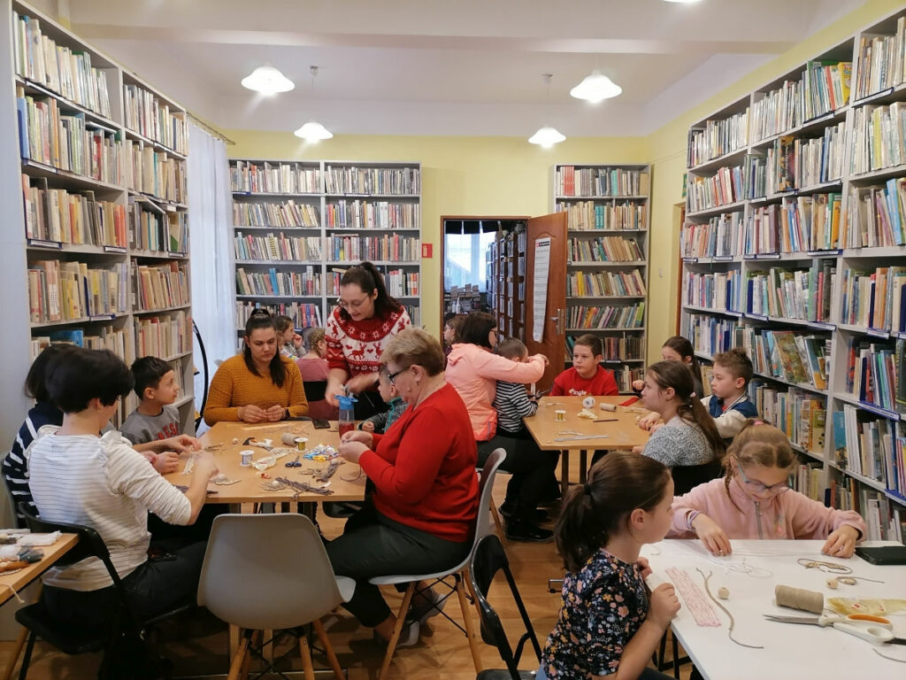 Na zdjęciu widocznych jest 6 osób dorosłych i 12 dzieci podczas wykonywania makramowych aniołków.