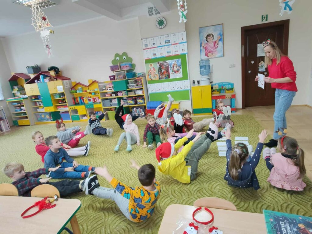 – Na zdjęciu widocznych jest 17 przedszkolaków i bibliotekarz wykonujących ćwiczenia gimnastyczne.