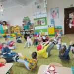– Na zdjęciu widocznych jest 17 przedszkolaków i bibliotekarz wykonujących ćwiczenia gimnastyczne.