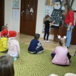 Na zdjęciu widocznych jest 8 przedszkolaków, 7 przedszkolaków siedzi na dywanie a 1 dziewczynka losuje zadanie od Mikołaja