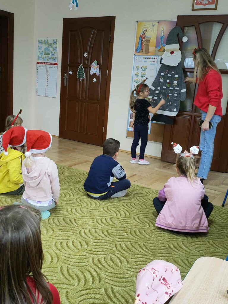 Na zdjęciu widocznych jest 8 przedszkolaków, 7 przedszkolaków siedzi na dywanie a 1 dziewczynka losuje zadanie od Mikołaja