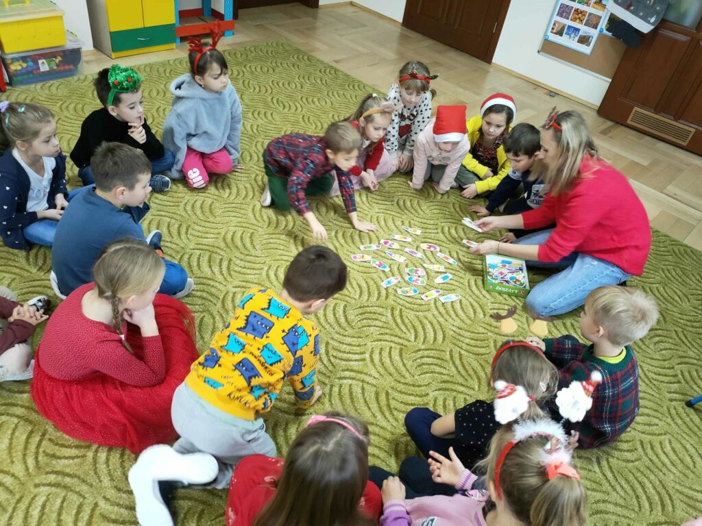 Na zdjęciu widocznych jest 17 przedszkolaków i bibliotekarz podczas grania w grę planszową.