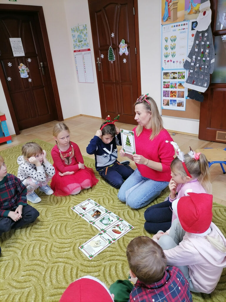 Na zdjęciu widoczny jest bibliotekarz trzymający karty z tradycjami bożonarodzeniowymi oraz 8 przedszkolaków siedzących na dywanie.