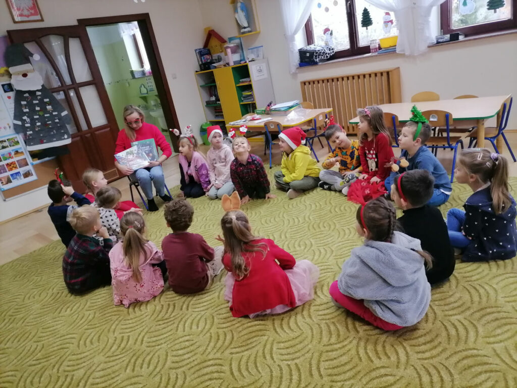 Na zdjęciu widoczny jest bibliotekarz trzymający 2 książki oraz 17 przedszkolaków (10 dziewczynek i 7 chłopców) siedzących na dywanie.