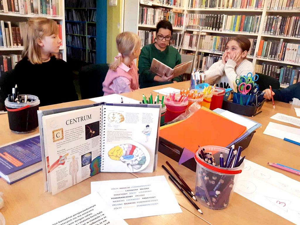 na zdjęciu widać salę biblioteczną oraz dzieci siedzące przy stole i słuchające czytaną im książkę