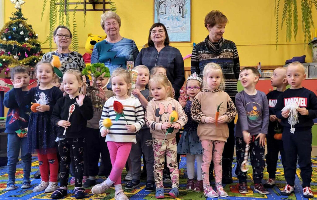 na zdjęciu widać dzieci i seniorki stojące w sali przedszkolnej