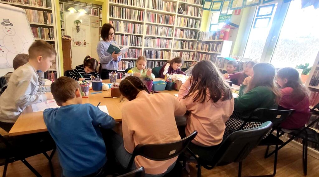 na zdjęciu widać dzieci siedzące wokół stołu w sali bibliotecznej i tworzące prace plastyczną