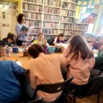 na zdjęciu widać dzieci siedzące wokół stołu w sali bibliotecznej i tworzące prace plastyczną