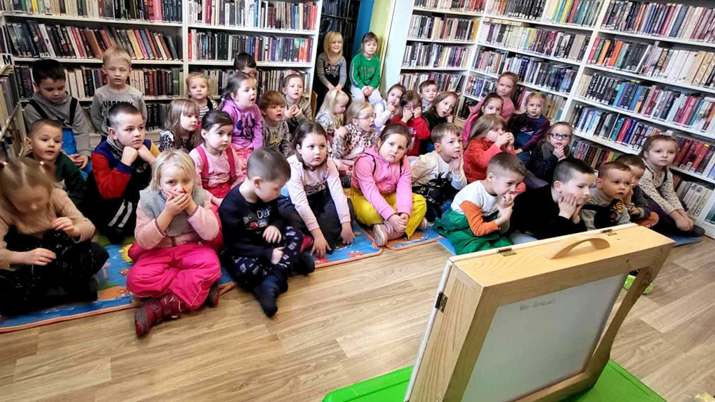 na zdjęciu widać dzieci w wieku przedszkolnym siedzące na podłodze w sali bibliotecznej