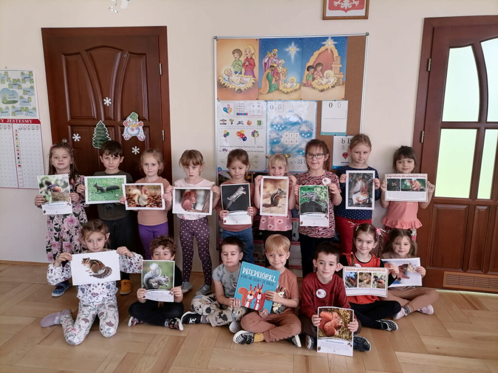 – Na zdjęciu widoczna jest grupa 16 przedszkolaków (11 dziewczynek i 5 chłopców) pozujących do wspólnego zdjęcia. 9 przedszkolaków stoi a 7 siedzi na podłodze, 2 chłopców trzyma książkę, a pozostałe dzieci trzymają zdjęcia wiewiórek.