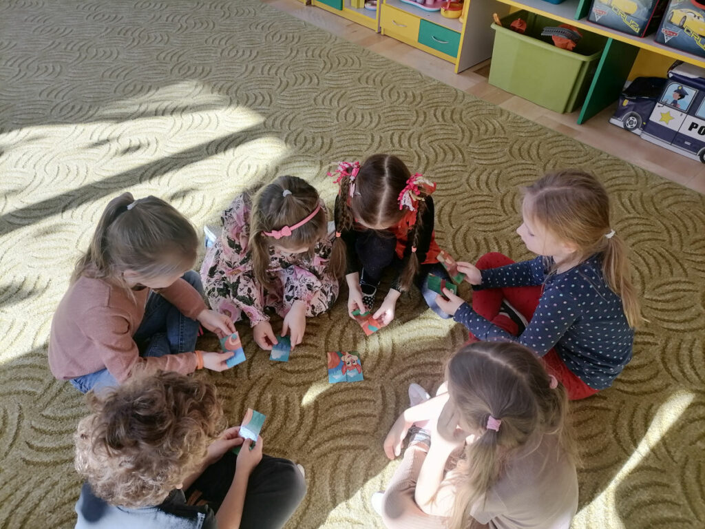 Na zdjęciu widoczna jest grupka 6 przedszkolaków siedzących na dywanie i układających puzzle