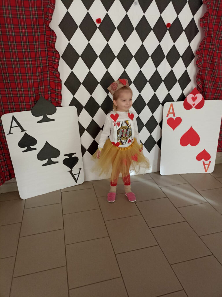 Na zdjęciu widoczna jest dziewczynka w stroju kart pozująca na ściance.