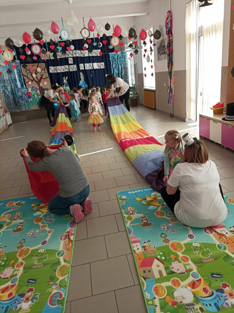 Na zdjęciu widoczna jest grupa przedszkolaków biorących udział w zabawie wyścigi w króliczych norkach czyli tunelach animacyjnych. Dzieci przechodzą przez tunele animacyjne trzymane przez panie. W tle dekoracje tematyczne.