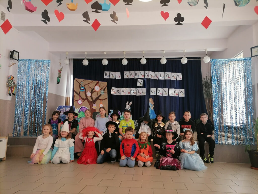 Na zdjęciu widoczna jest grupa 19 dzieci, 8 dzieci siedzi na podłodze a 11 dzieci siedzi na scenie. Dzieci pozują do wspólnego zdjęcia. W tle dekoracje tematyczne.