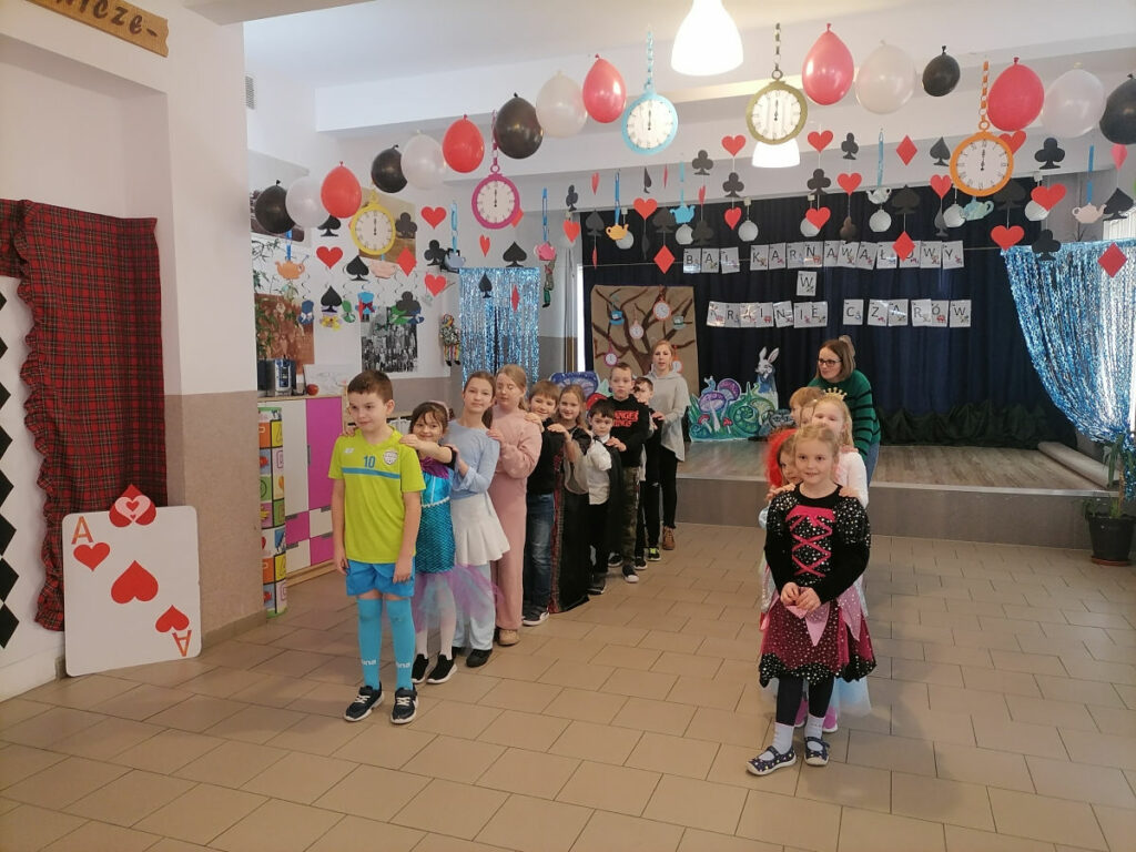 – Na zdjęciu widoczna jest grupa dzieci i dwie wychowawczynie, ustawionych w dwóch rzędach. W tle dekoracje tematyczne.