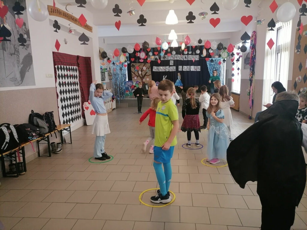 na zdjęciu widoczna jest grupa dzieci biorących udział w zabawie skoki do króliczej norki, polegającej na tym, że na przerwę w muzyce należy jak najszybciej wskoczyć do plastikowej obręczy znajdującej się na podłodze. Po każdej przerwie w muzyce jedna obręcz jest zabierana, a dziecko które nie zdążyło wskoczyć do wolnej obręczy odpada z gry.