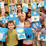 Na zdjęciu widać dzieci w sali bibliotecznej trzymające kolorowe prace plastyczne
