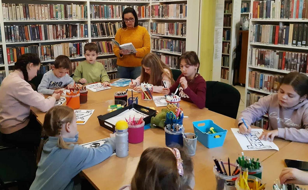 na zdjęciu widać dzieci siedzące i przy stole i słuchające czytająca im bibliotekarkę