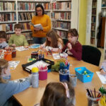 na zdjęciu widać dzieci siedzące i przy stole i słuchające czytająca im bibliotekarkę
