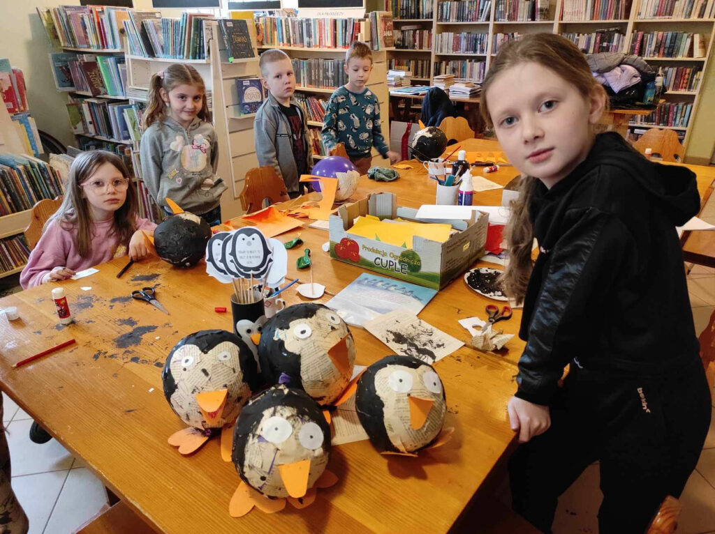 na zdjęciu widać salę biblioteczną , na środku pomieszczenia stoi stół, przy stole siedzą dzieci , wykonują pracę plastyczną